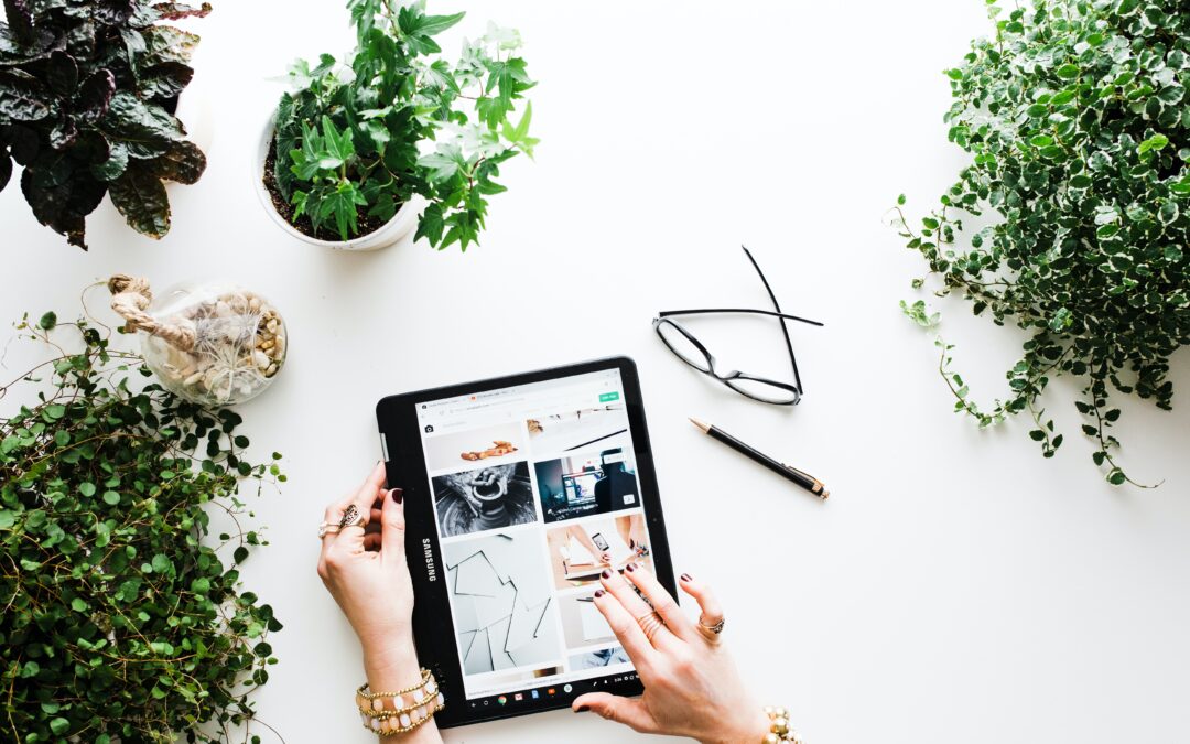 Flat lay image of a woman on a tablet, with glasses, a pen and lots of plants on a white desk