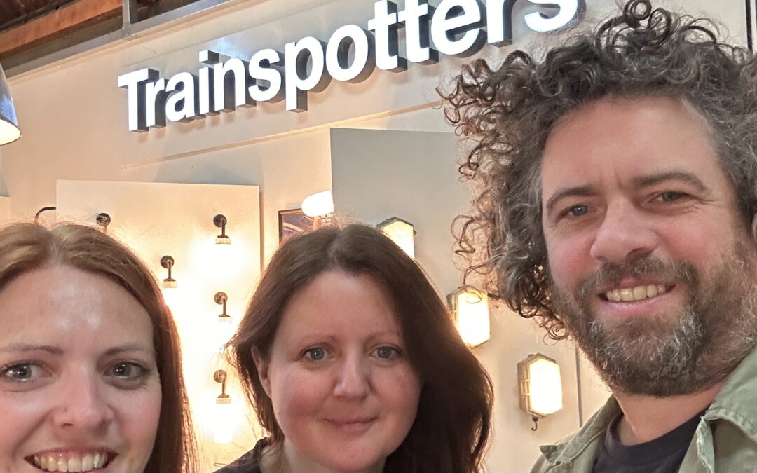 Two women and a man smiling and stood infront of a white 'trainspotters' sign